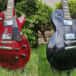 Dvě fotografie elektrických kytar typu Les Paul, jedna v červené a druhá v černé barvě. Obě mají dva humbuckery, tmavý hmatník a černý plechtrpickgua…