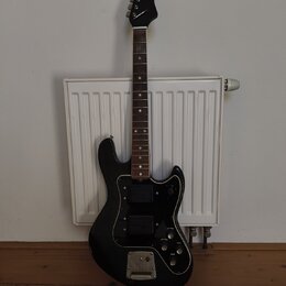 Černá elektrická kytara s černým pickguardem a hnedým hmatníkem. Na hlavě je bílý nápis "SAGA". Nástroj má dva snímače typu humbucker, tři ovládací k…