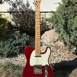 Elektrická kytara typu Telecaster v červené barvě s bílým pickguardem a světle hnědým javorovým hmatníkem. Na hlavě je viditelné logo Fender. Povrch…