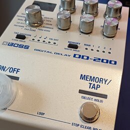 Bílý efekt pedál s nápisy BOSS DIGITAL DELAY DD-200. Na horní části jsou ovládací knoflíky a přepínače s popisy funkcí, jako je TAP DIVISION nebo MEM…