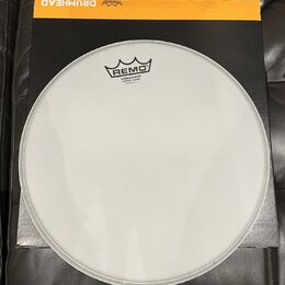 Bílá kruhová bubenová kožíška položená na černém kartonu s žlutým horním okrajem. Na kožíšku je černé logo REMO a nápisy AMBASSADOR VINTAGE COATED MA…