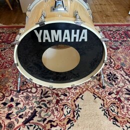 Světle béžový basový buben s černým předním koženem. Na koženě je bílý nápis YAMAHA a uprostřed je vyříznutý kruhový otvor. Nástroj má chromované lug…