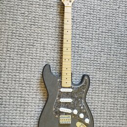 Černá elektrická kytara s tvarem typu Stratocaster a tři bílými snímači. Nástroj má světlý dřevěný hmatník a plech s tmavým mramorovaným vzorem. Na h…
