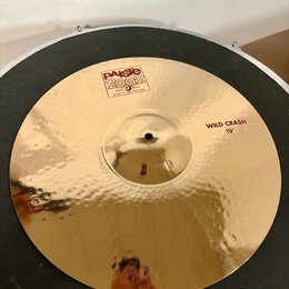 Zlatý kovový činel s kladeným povrchem. Na povrchu jsou červené nápisy „Paiste 2002“, „TRADE MARK MADE IN SWITZERLAND“ a „WILD CRASH 19"“. Povrch je…