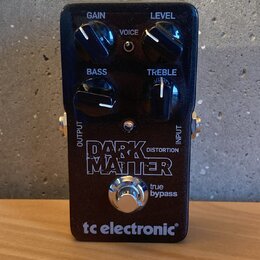Černý efekt pedál s nápisy GAIN, LEVEL, BASS a TREBLE u ovládacích knoflíků a přepínačem VOICE. Uprostřed je velký bílý nápis DARK MATTER DISTORTION…