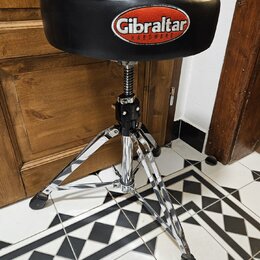 Černý polstrovaný bubenícký židun s chromovaným třínohým stativem. Na sedadle je bílo-červené logo s nápisem Gibraltar HARDWARE. Na černé části mecha…