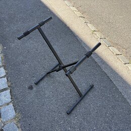 Černý kovový stojan na hudební nástroj s dvěma polstrovanými oporami. Stojan má sklapovací konstrukci a stojí na asfaltovém povrchu. Na fotce nejsou…