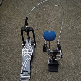 Černý basový pedál s kovovým šlapadlem, na kterém je bílý nápis DIXON. K pedálu vede flexibilní kovové táhlo k mechanismu u bubnu. Mechanismus je vyb…