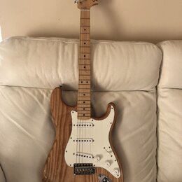Elektrická kytara s přírodním dřevěným povrchem těla a hmatníkem. Na hlavě je viditelné logo Fender Stratocaster. Bílý pickguard obsahuje tři snímače…