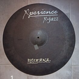 Kulatý činel s tmavým, matným povrchem. Na horní části je bílý nápis „Xperience“ a pod ním „X-jazz“, vlevo od něj je menší nečitelný text. Ve spodní…