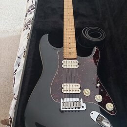 Černá elektrická kytara s tvarem typu Stratocaster, světlým hmatníkem a červeně vzorovaným pickguardem. Nástroj má tři snímače, tři ovládací knofíky…