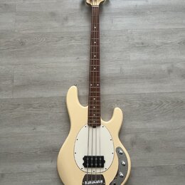 Elektrická basová kytara v krémové barvě s bílým pickguardem. Na hlavě hmatníku je viditelné logo „Sterling by Music Man“. Nástroj má hnědý hmatník,…