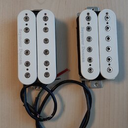 Dvě elektrické kytarové snímače typu humbucker s bílými plastovými kryty. Na obou je z levé strany svisle napsán nápis Seymour Duncan. Snímače mají č…