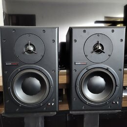 Dva černé regálové reproduktory umístěné na černých stojanech. Na přední straně každého reproduktoru je nápis „dynaudio acoustics“ na červeném pozadí…