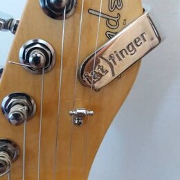 Detail hlavy elektrické kytary z přírodního dřeva. Na dřevě je viditelné částečné logo Fender. Na strunách je upevněn kovový stahovací kousek s nápis…