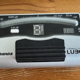 Stříbrný chromatický tuner s černým gumovým pedálem. Na zařízení jsou nápisy Ibanez, CHROMATIC TUNER a LUBO. Horní část obsahuje displej s označením…