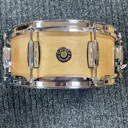 Fotografie znázorňuje snare drum s dřevěným korpusem v přírodní světle barvě a chromovaným obručemi. Uprostřed korpusu je umístěno kulaté logo s nápi…