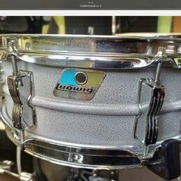 Stříbrný snare drum s texturovaným povrchem a chromovanými obručemi. Na korpusu je viditelný štítek s logem ludwig a nečitelným nápisem pod ním. Nást…