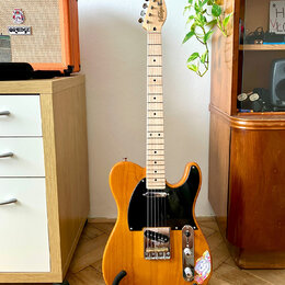 Elektrická kytara typu Telecaster v žluté barvě s černým pickguardem a světlým hnedým dřevěným hmatníkem. Na hlavě je vidět nápis "Squier" a pod ním…