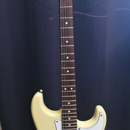 Elektrická kytara s krémovým korpusem, bílým pickguardem a hnedou hmatníkovou deskou. Na hlavě je černý nápis „Fender STRATOCASTER“. Nástroj má tři s…