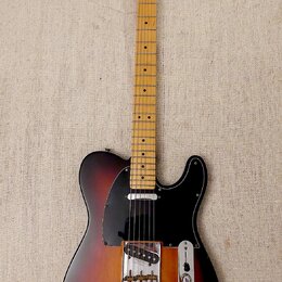 Elektrická kytara s tmelelovým povrchem v přechodu od černé přes červenou k žluté. Na hlavě hmatníku je nápis Fender TELECASTER. Kytara má černý pick…