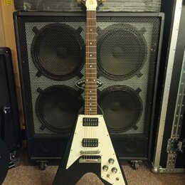 Černá elektrická kytara s bílým pickguardem a Flying V tvarem těla. Na hlavě je viditelné logo "Gibson". Na spodní části těla je nápis "Epiphone". Ky…