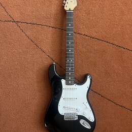 Černá elektrická kytara s bílým pickguardem a hnedým hmatníkem. Na hlavě je viditelné logo Fender. Nástroj má tři snímače typu single-coil a chromova…