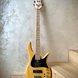 Elektrická basová kytara s přírodním dřevěným povrchem těla a hmatníkem. Na hlavě je viditelné logo ve tvaru motýla. Instrument má černý pickguard, č…
