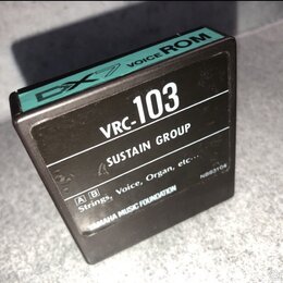 Černý čtvercový modul s nápisem VRC-103 a textem SUSTAIN GROUP. Horní hrana je tyrkysově zelená s nápisem DX7 VOICE ROM. Dole jsou uvedeny kategorie…