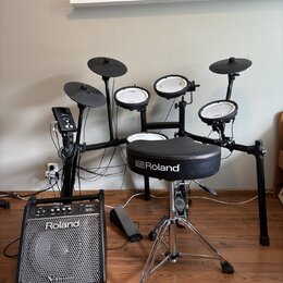 Elektronická bicí souprava s černým rámem, několika pady a činely. Na sedáku je bílý nápis Roland a na př bundled kombu v popředí nápisy Roland a V-D…