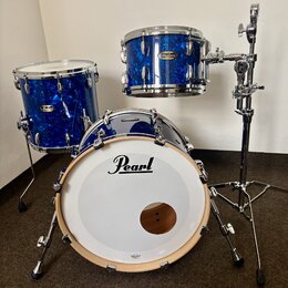 Modrá bubnová sada s lesklým povrchem a chromovaným kováním. Na basovém bubnu je bílý kožený přední plát s černým nápisem Pearl. Na tomu a tom-tomu j…