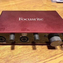 Červená audio rozhraní s nápisem Focusrite na horní straně. Na předním panelu jsou dva kombinované vstupy, dva ovládací knofíky zyska a velký knofík…