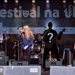 Fotografie zachycuje hudební kapelu na pódiu před černým bannerem s nápisem „Festival na ulici MULTIŽÁCROVÝ FESTIVAL V ULICÍCH PLZNĚ“. Vlevo stojí ky…