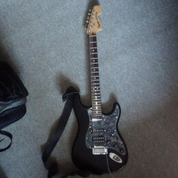 Černá elektrická kytara s tvarem typu Stratocaster a hmatníkem z tmavého dřeva. Na hlavě je viditelné logo "Squier". Kytara má stříbrný pickguard s t…