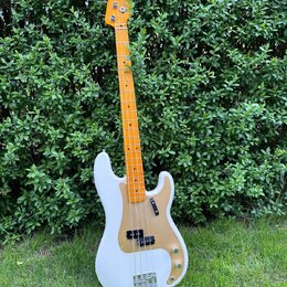 Bílá elektrická basová kytara s hnědým javorovým hmatníkem a zlatým plechem. Na hlavě nástroje je viditelné logo Fender. Instrument má dva snímače, z…
