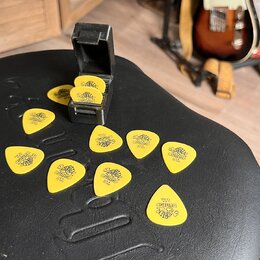 Žluté kytarové trsátka s černým potiskem loga „dunlop“ a nápisem „U.S.A.“. Na některých kusech je viditelný nápis „.73 mm“. Trsátka jsou rozprostřena…