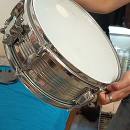Fotografie zachycuje snare drum s chromovaným kovovým korpusem a bílým membránou. Nástroj drží osoba v rukou. Na viditelných částech nástroje nejsou…