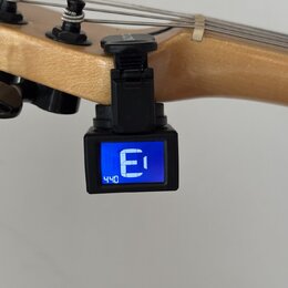 Černý digitální ladicí přístroj s modrým displejem, na kterém je zobrazeno písmeno „E“ a číslo „440“. Ladička je pomocí svorky připevněna na hmatník…
