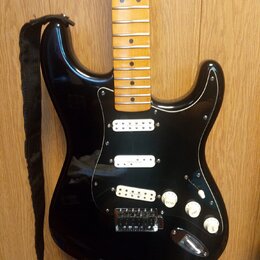 Černá elektrická kytara s tvarem typu Stratocaster a černým pickguardem. Má světle hnědý javorový hmatník a tři bílé snímače. Na pickguardu je vidite…