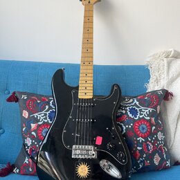 Černá elektrická kytara s mírně prohnutým tělem a světle hnědým hnedem hmatníkem. Na hlavě je nápis Squier Stratocaster. Tělo má černý pickguard, tři…
