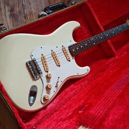 Bílá elektrická kytara typu Stratocaster s bílým pickguardem a hnedým hmatníkem, umístěná v otevřeném kufru s červeným polstrováním. Na hlavě hmatník…