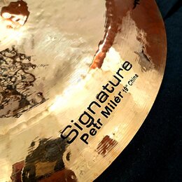 Detailní záběr na povrch mosazného činele s kovaným povrchem. Na čineli je černý nápis „Signature“ a pod ním menší text „Petr Milér 19" China“. Povrc…