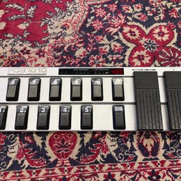 Stříbrný MIDI pedál s deseti černými spínači a dvěma expresními pedály v dolní části. Na pravém boku je svisle napsáno FCB1010 MIDI FOOT CONTROLLER.…