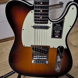 Elektrická kytara typu Telecaster s barevným přechodem korálovo-černé (sunburst) a krémovým pickguardem. Na pickguardu je černý štítek s nápisy „Fend…