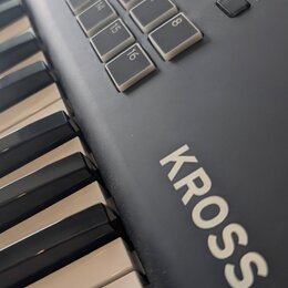 Detailní pohled na černý digitální klavír s bílými klávesami. Na korpusu je výrazný bílý nápis KROSS. Horní panel obsahuje sadu bílých čtvercových tl…