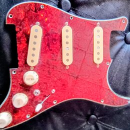 Červený pickguard s texturou připomínající mramor nebo plameny a stříbrným okrajem. Je na něm osazeno tři bílé snímače s viditelnými póly. V dolní čá…