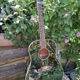 Černá akustická kytara s lesklým povrchem a hnědým dřevěným hmatníkem. Na hlavě nástroje je bílý nápis Gibson. Kytara je umístěna na stojanu před zel…