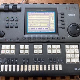 Šedý music sequencer značky YAMAHA, model QY700. Zařízení má v horní části displej s modrým podsvícením a pod ním řadu ovládacích tlačítek. V pravé č…