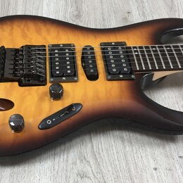 Elektrická kytara s prohýbaným topem v žlutě-hnědém sunburst provedení s viditnou strukturou dřeva. Nástroj má tři snímače (dvě humbuckery a jeden si…