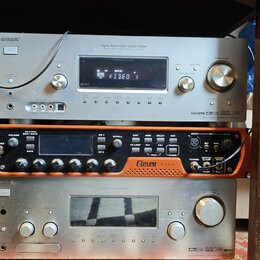 Tři stohované elektronické komponenty v regálu. Horní je stříbrný receiver s nápisy SONY, Digital Audio/Video Control Center a HDMI. Prostřední je or…